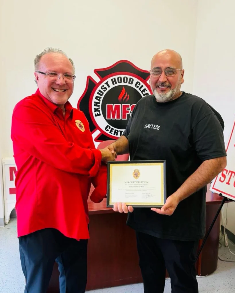 Two men smiling and shaking hands; one is holding a framed certificate. They are standing in front of a wall with a logo that says "Exhaust Hood Cleaning MFS Certified.